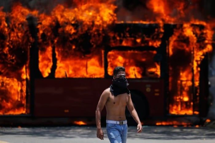 Một người biểu tình chống chính phủ đi bộ gần một chiếc xe buýt bị đốt cháy trong các cuộc đụng độ ở Caracas, Venezuela năm 2019. Ảnh: AP.