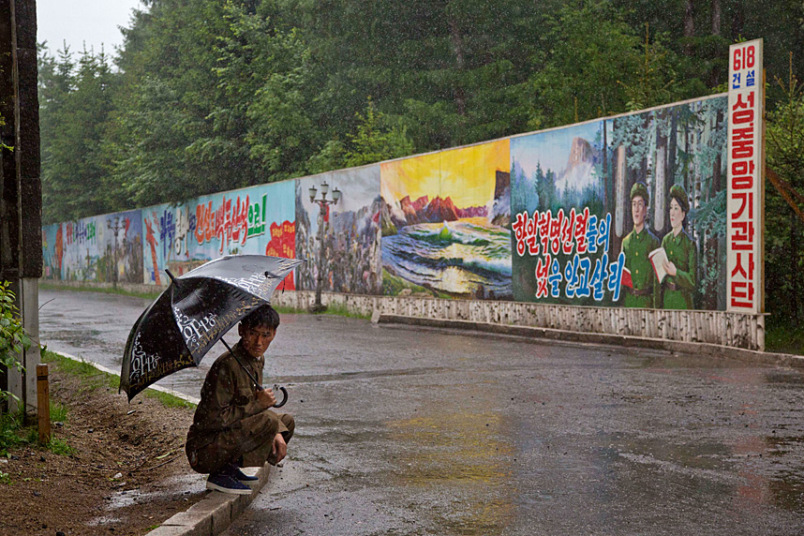 Người đàn ông ngồi trú mưa cạnh những khẩu hiệu lớn tại thị trấn Samjiyon.