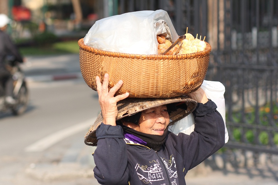 Một người phụ nữ trung niên bán bánh dạo, đội trên đầu rất nhiều các loại bánh như bánh rán, bánh mì...