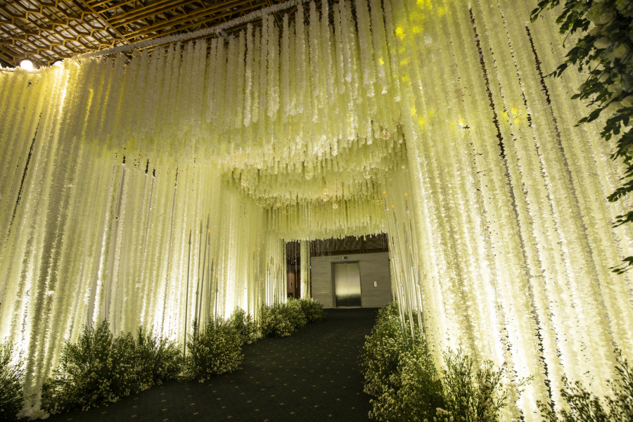 Dọc lối đi vào khu vực sảnh tiệc được bao phủ bằng những dây hoa trắng muốt thả từ trần nhà xuống. Bên dưới là hàng nghìn bông hoa thạch thảo làm đẹp cho lối đi.